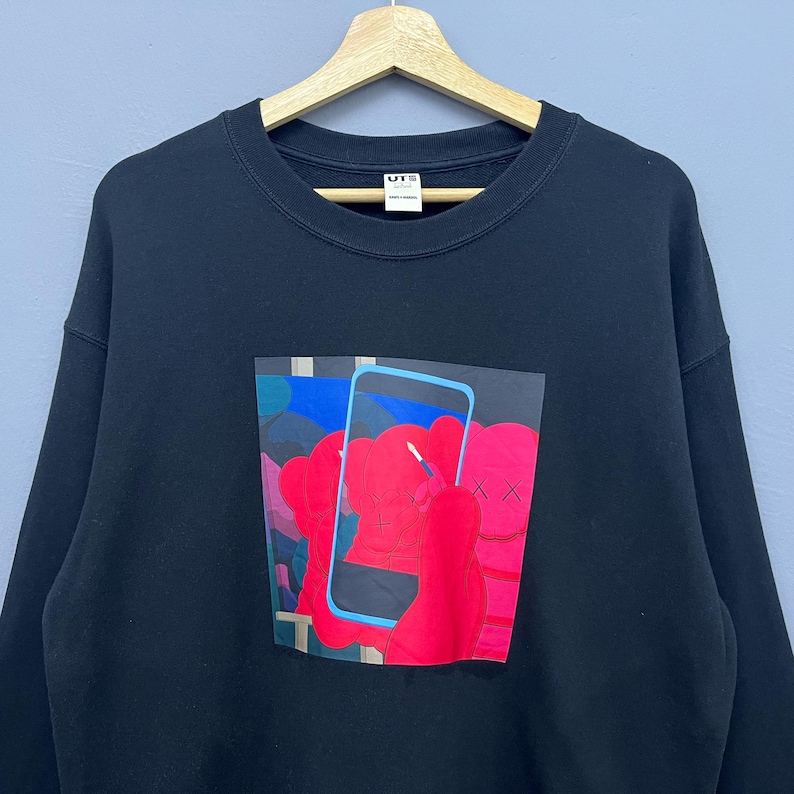 May include: A black crewneck sweatshirt featuring a graphic print on the chest. The design depicts a bright red cartoon character with X-shaped eyes holding a light blue smartphone. The garment has a ribbed neckline and a white interior brand label.