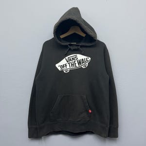 Op de afbeelding: Zwarte hoodie met een kangoeroezak en het witte Vans-logo. Het logo toont de tekst "Vans Off The Wall" op een skateboard. Een klein rood label is zichtbaar op de zak.