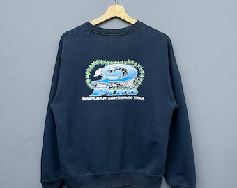 Può includere: Felpa blu navy scuro con girocollo. Il retro presenta una grafica di un'onda con la scritta "PIKO" in blu e bianco, circondata da un motivo floreale giallo e blu. Il testo "HAWAIIAN LONGBOARD WEAR" è sottostante.