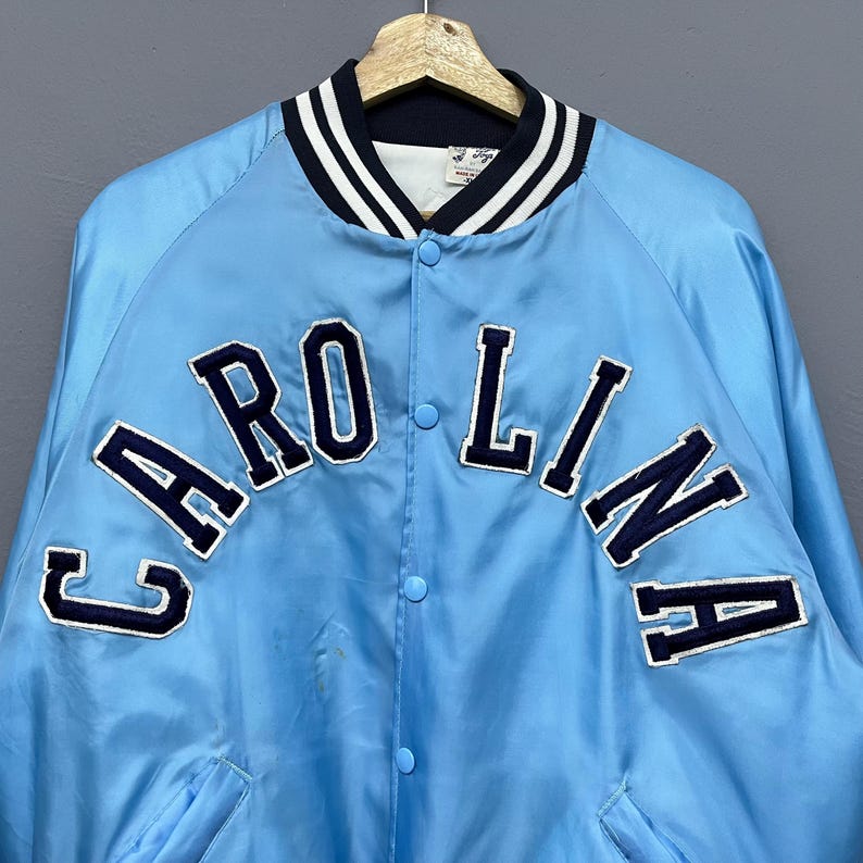 May include: Light blue satin jacket with the word "CAROLINA" in navy blue block letters across the chest. The jacket has a navy blue and white striped collar and cuffs. The jacket is buttoned up and hanging on a wooden hanger.