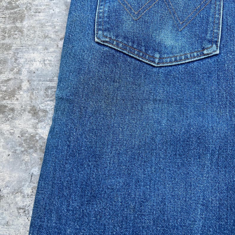 May include: Close-up of a section of blue denim jeans. The image shows the texture of the fabric and a back pocket with a decorative stitch design. The jeans are a medium wash.