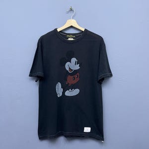 May include: Black t-shirt with a Mickey Mouse graphic. The Disney Resort logo is visible at the top. The shirt is hanging on a wooden hanger. The t-shirt is a size L.