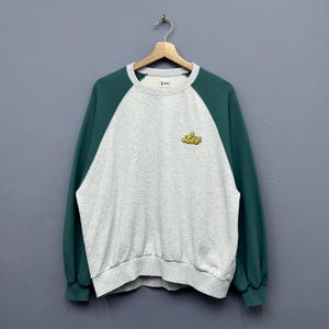 Op de afbeelding: Een grijze sweatshirt met donkergroene raglanmouwen. Het woord "Lee" is in het geel op de borst geborduurd. De halslijn en manchetten zijn grijs.