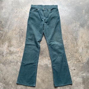 May include: Teal corduroy trousers with a classic five-pocket design. The trousers have a button and zip closure and a flared leg. The corduroy fabric has vertical ridges and a soft texture. The trousers are laid flat on a concrete surface.