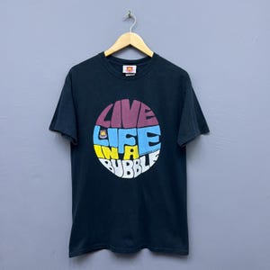 Puede incluir: Camiseta negra con la frase "LIVE LIFE IN A BUBBLE" en un diseño circular colorido. El texto está en una fuente retro, con colores morado, azul, amarillo y blanco. La camiseta está colgada en una percha de madera.