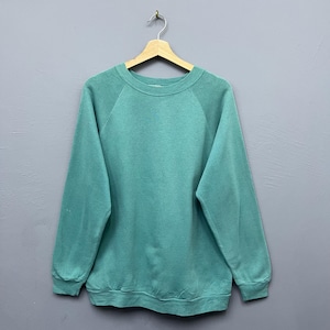Könnte beinhalten: Ein teal-farbenes Sweatshirt mit Rundhalsausschnitt und langen Ärmeln, das an einem Holzbügel hängt. Das Kleidungsstück hat ein leicht verwaschenes Aussehen, was darauf hindeutet, dass es Vintage sein könnte. Der Hintergrund ist einfarbig grau.
