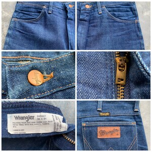 K&ouml;nnte beinhalten: Nahaufnahmen von dunkelblauen Wrangler Jeans. Details sind ein Knopf mit dem Wrangler-Logo, ein Messingrei&szlig;verschluss und ein Lederaufn&auml;her auf der Ges&auml;&szlig;tasche. Das Etikett lautet "945NAY" und "34 x 34".