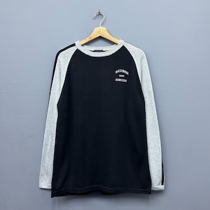 Op de afbeelding: Zwart en grijs Reebok atletisch sweatshirt op een houten hanger. Het sweatshirt heeft grijze mouwen en een zwarte body. De tekst "REEBOK 1895 ATHLETIC" is op de bovenborst gedrukt.