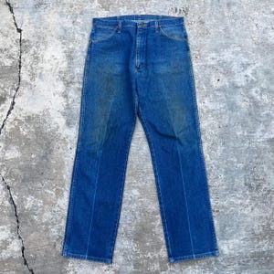 May include: A pair of blue denim jeans with a classic five-pocket design. The jeans have a button and zipper closure, belt loops, and a faded appearance. The jeans are laid flat on a concrete surface.