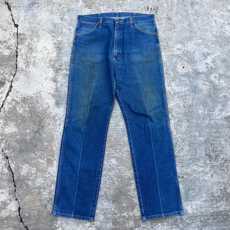 May include: A pair of blue denim jeans with a classic five-pocket design. The jeans have a button and zipper closure, belt loops, and a faded appearance. The jeans are laid flat on a concrete surface.