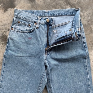 K&ouml;nnte beinhalten: Hellblaue Jeans mit teilweise ge&ouml;ffnetem Rei&szlig;verschluss. Die Jeans haben ein klassisches F&uuml;nf-Taschen-Design mit sichtbaren Nieten und einem Knopfverschluss. Der Stoff weist Gebrauchsspuren auf.
