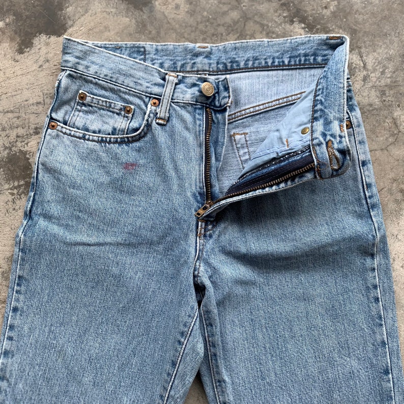 K&ouml;nnte beinhalten: Hellblaue Jeans mit teilweise ge&ouml;ffnetem Rei&szlig;verschluss. Die Jeans haben ein klassisches F&uuml;nf-Taschen-Design mit sichtbaren Nieten und einem Knopfverschluss. Der Stoff weist Gebrauchsspuren auf.