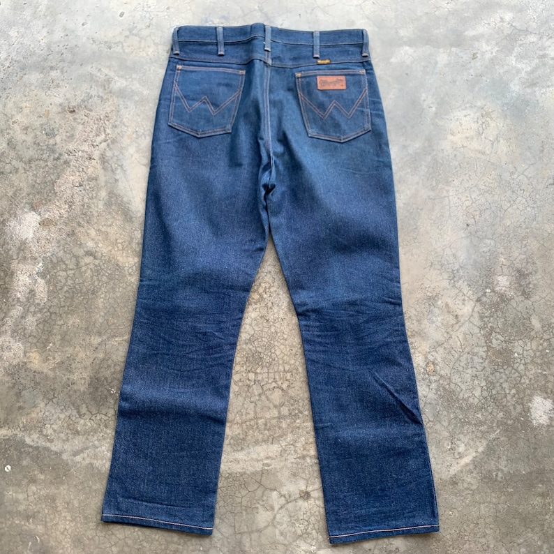 K&ouml;nnte beinhalten: Dunkelblaue Jeans aus Denim mit klassischem F&uuml;nf-Taschen-Design. Die Jeans haben einen braunen Lederaufn&auml;her mit dem Wrangler-Logo und charakteristischen N&auml;hten auf den Ges&auml;&szlig;taschen. Die Jeans liegen flach und zeigen die R&uuml;ckansicht.