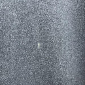 Op de afbeelding: Close-up van een donker marineblauwe stof met een kleine, lichtgekleurde vlek. De stof heeft een getextureerde uitstraling, met zichtbare vezels en een ietwat versleten look. De vlek is klein en onregelmatig van vorm.