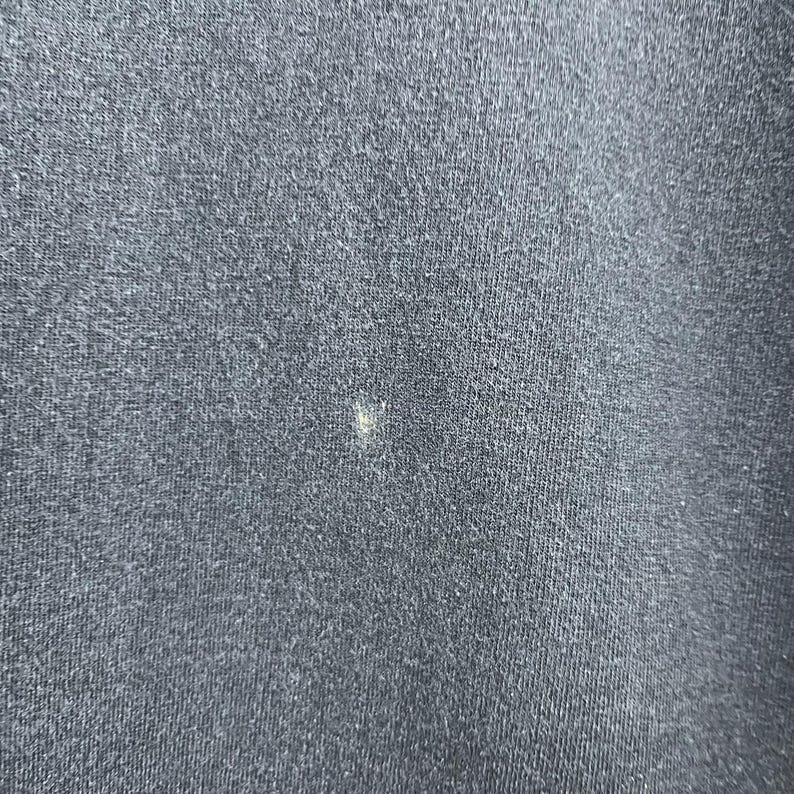 Op de afbeelding: Close-up van een donker marineblauwe stof met een kleine, lichtgekleurde vlek. De stof heeft een getextureerde uitstraling, met zichtbare vezels en een ietwat versleten look. De vlek is klein en onregelmatig van vorm.