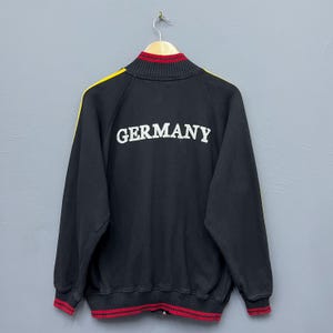 Könnte beinhalten: Schwarze Trainingsjacke mit Reißverschluss und dem Wort "GERMANY" in weißen Blockbuchstaben auf dem Rücken. Die Jacke hat gelbe Streifen an den Ärmeln und rot-gelb gestreifte Verzierungen an Kragen, Bündchen und Saum.