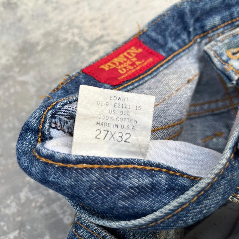 Pode incluir: Close-up de jeans de ganga azul com uma etiqueta vermelha Edwin. Os jeans t&ecirc;m uma etiqueta branca com o texto "EDWIN MADE IN U.S.A." e o tamanho 27x32. Os jeans s&atilde;o feitos de 100% algod&atilde;o.