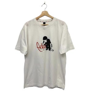 Puede incluir: Camiseta blanca con un gráfico negro de una persona arrodillada y el texto rojo "Acto function". La camiseta está colgada en una percha de madera.