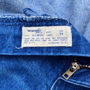 Pode incluir: Close-up de jeans azuis com uma etiqueta da marca Wrangler. A etiqueta mostra o tamanho da cintura como 89 cm e o comprimento como 86 cm. O z&iacute;per &eacute; prateado. A etiqueta tamb&eacute;m inclui instru&ccedil;&otilde;es de lavagem.