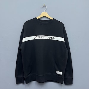 May include: Black crew neck sweatshirt with a white horizontal stripe across the chest. The text "DCSHOECOUSA" is printed in black on the white stripe. A small white tag is on the lower right side of the sweatshirt.