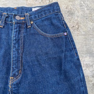 K&ouml;nnte beinhalten: Nahaufnahme einer dunkelblauen Jeans. Die Jeans hat einen Knopfverschluss, Vordertaschen und orangefarbene N&auml;hte. Die Jeans hat eine lila Niete an der Vordertasche. Das Wort "AUTHENTIC" ist auf dem Bund zu sehen.