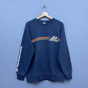 Puede incluir: Una sudadera azul marino de manga larga con cuello redondo y puños acanalados. El pecho muestra un gráfico con rayas horizontales naranjas, amarillas y blancas, el logotipo de Piko y el texto Hawaiian Longboard Wear. La manga también tiene texto.