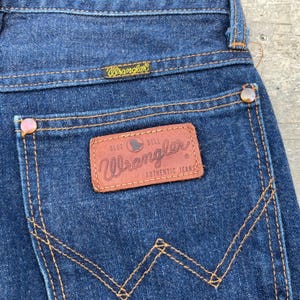 K&ouml;nnte beinhalten: Nahaufnahme einer dunkelblauen Jeans mit einem braunen Lederaufn&auml;her auf der Ges&auml;&szlig;tasche. Der Aufn&auml;her zeigt das Wrangler-Logo und die Worte "Blue Bell Authentic Jeans". Die Jeans hat kupferfarbene Nieten und N&auml;hte.