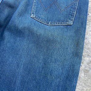 May include: Close-up of a section of blue denim jeans. The image highlights the texture of the fabric and a back pocket with a stitched design. The denim has a faded appearance, suggesting wear.