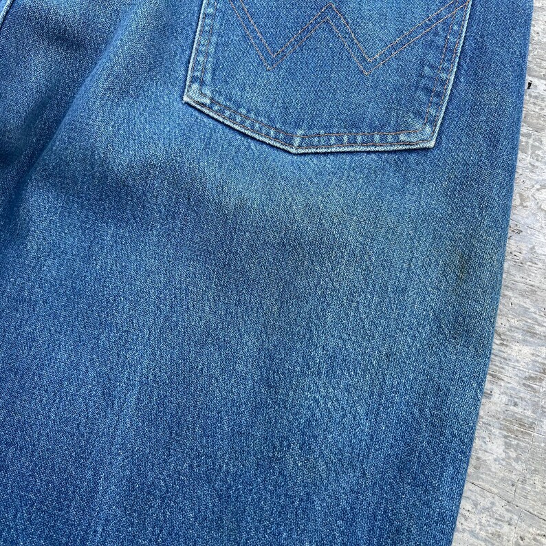 May include: Close-up of a section of blue denim jeans. The image highlights the texture of the fabric and a back pocket with a stitched design. The denim has a faded appearance, suggesting wear.