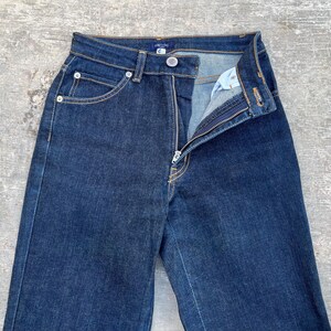 May include: Dark wash denim jeans with a high waist and a zipper fly. The jeans have classic five-pocket styling with silver rivets and a button closure. The fabric appears to be a medium-weight denim.