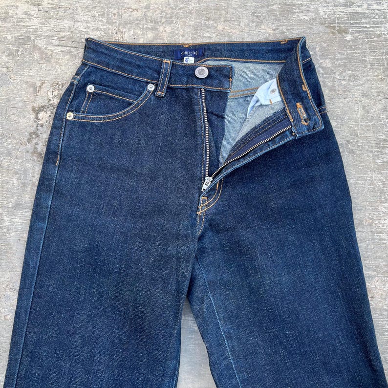 May include: Dark wash denim jeans with a high waist and a zipper fly. The jeans have classic five-pocket styling with silver rivets and a button closure. The fabric appears to be a medium-weight denim.