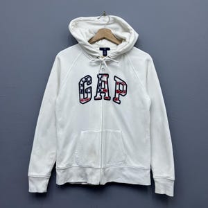 May include: White zip-up hoodie with a hood and a front pocket. The word "GAP" is displayed on the chest in a patriotic design with red, white, and blue colours. The hoodie is hanging on a wooden hanger.