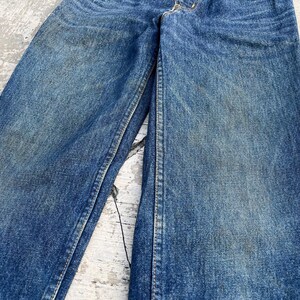 Pode incluir: Close-up de cal&ccedil;as de ganga azuis. As cal&ccedil;as de ganga mostram sinais de desgaste e desbotamento, com costuras vis&iacute;veis e um design cl&aacute;ssico de cinco bolsos. O denim tem uma apar&ecirc;ncia ligeiramente desgastada.