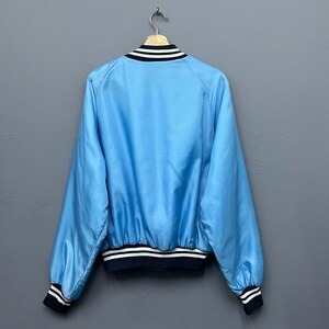 May include: Light blue bomber jacket with black and white striped trim on the collar, cuffs, and hem. The jacket is hanging on a wooden hanger against a gray background. The jacket has a shiny, satin-like appearance.