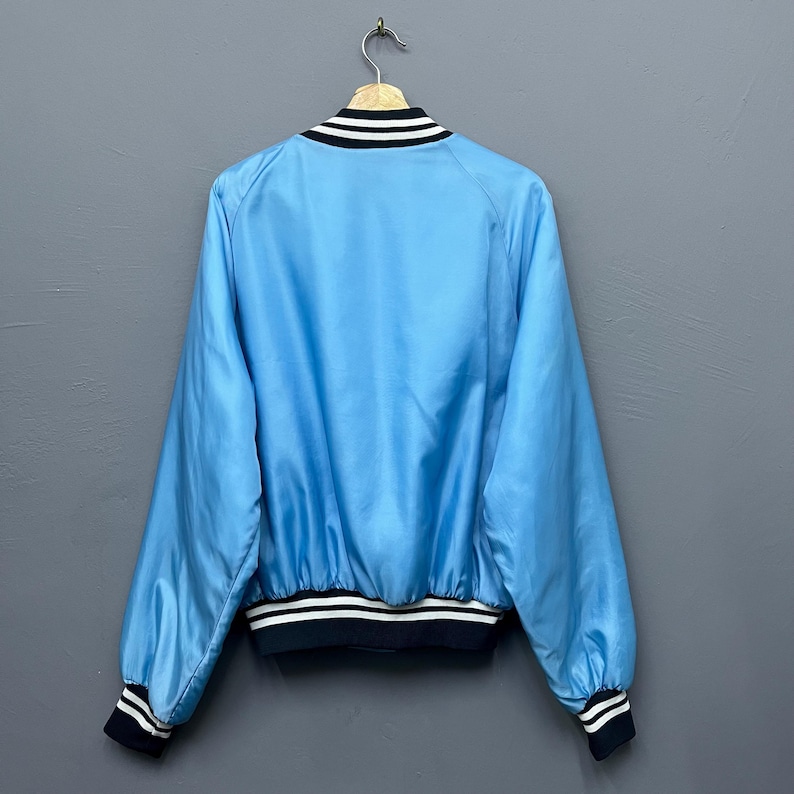 May include: Light blue bomber jacket with black and white striped trim on the collar, cuffs, and hem. The jacket is hanging on a wooden hanger against a gray background. The jacket has a shiny, satin-like appearance.