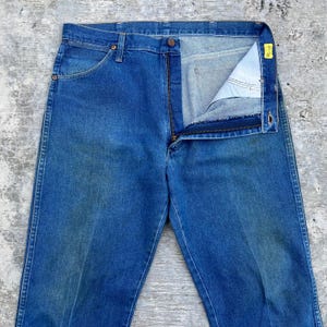 May include: Blue denim jeans with a partially unzipped fly. The jeans have a classic five-pocket design, with a button and zipper closure. The denim has a faded appearance, suggesting wear. The jeans are laid flat on a textured surface.