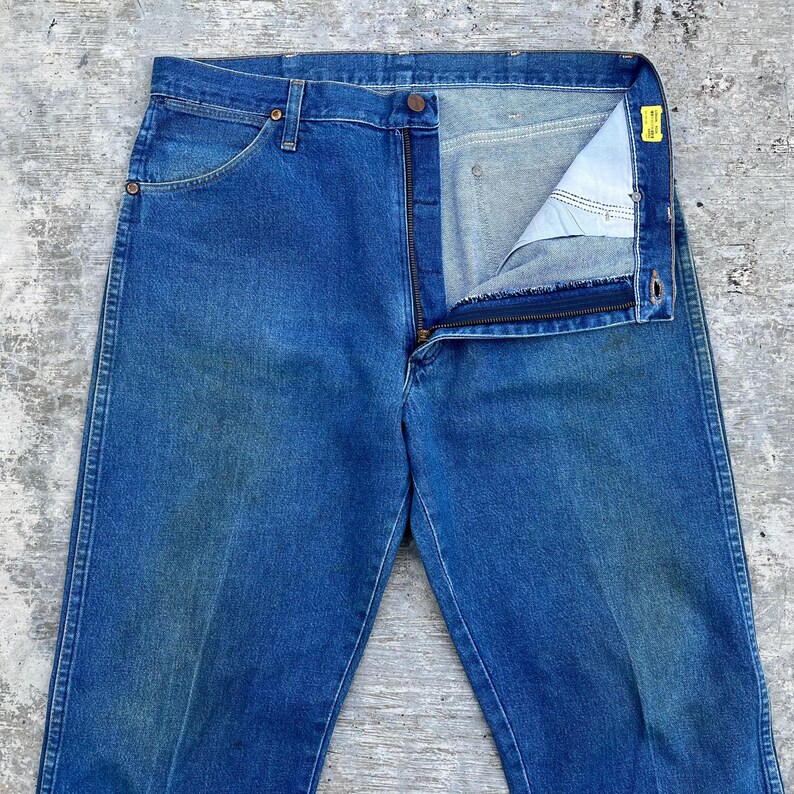 May include: Blue denim jeans with a partially unzipped fly. The jeans have a classic five-pocket design, with a button and zipper closure. The denim has a faded appearance, suggesting wear. The jeans are laid flat on a textured surface.