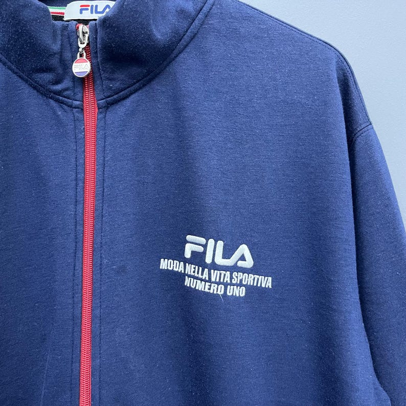 Puede incluir: Chaqueta azul marino con cremallera con el logo y el texto FILA. La chaqueta tiene una cremallera roja y un peque&ntilde;o logo FILA en el cuello. El texto de la chaqueta dice "MODA NELLA VITA SPORTIVA NUMERO UNO".