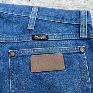 May include: Close-up of blue denim jeans with the Wrangler brand label on the back pocket. The jeans have a classic five-pocket design with copper-colored rivets and stitching. The Wrangler logo is also visible on a small black label.