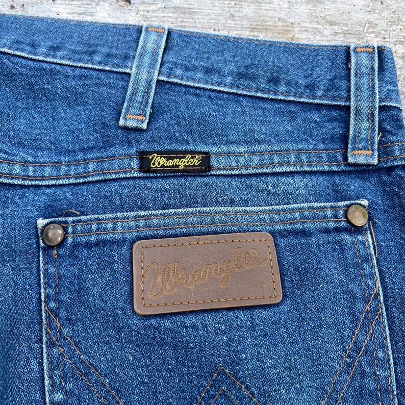 May include: Close-up of blue denim jeans with the Wrangler brand label on the back pocket. The jeans have a classic five-pocket design with copper-colored rivets and stitching. The Wrangler logo is also visible on a small black label.