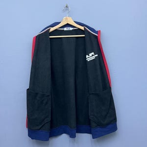 Puede incluir: Chaqueta negra con cremallera, cuello y pu&ntilde;os azul marino y rayas rojas en los laterales. La chaqueta tiene dos bolsillos delanteros y la palabra "AIR" impresa en blanco en el pecho. La chaqueta est&aacute; colgada en una percha de madera.