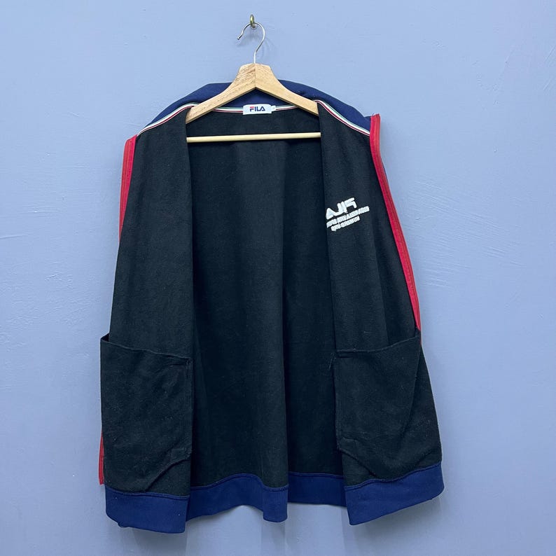 Puede incluir: Chaqueta negra con cremallera, cuello y pu&ntilde;os azul marino y rayas rojas en los laterales. La chaqueta tiene dos bolsillos delanteros y la palabra "AIR" impresa en blanco en el pecho. La chaqueta est&aacute; colgada en una percha de madera.