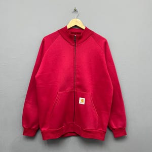 Könnte beinhalten: Ein leuchtend rotes Sweatshirt mit Reißverschluss, Stehkragen, langen Ärmeln mit gerippten Bündchen und einem durchgehenden schwarzen Frontreißverschluss. Ein kleines rechteckiges Carhartt-Markenetikett ist auf der Vordertasche aufgenäht.