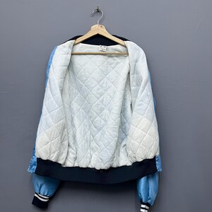 May include: A vintage satin bomber jacket in light blue and black, hanging on a wooden hanger. The jacket features a quilted white lining and black ribbed cuffs and hem. The sleeves have a light blue stripe.