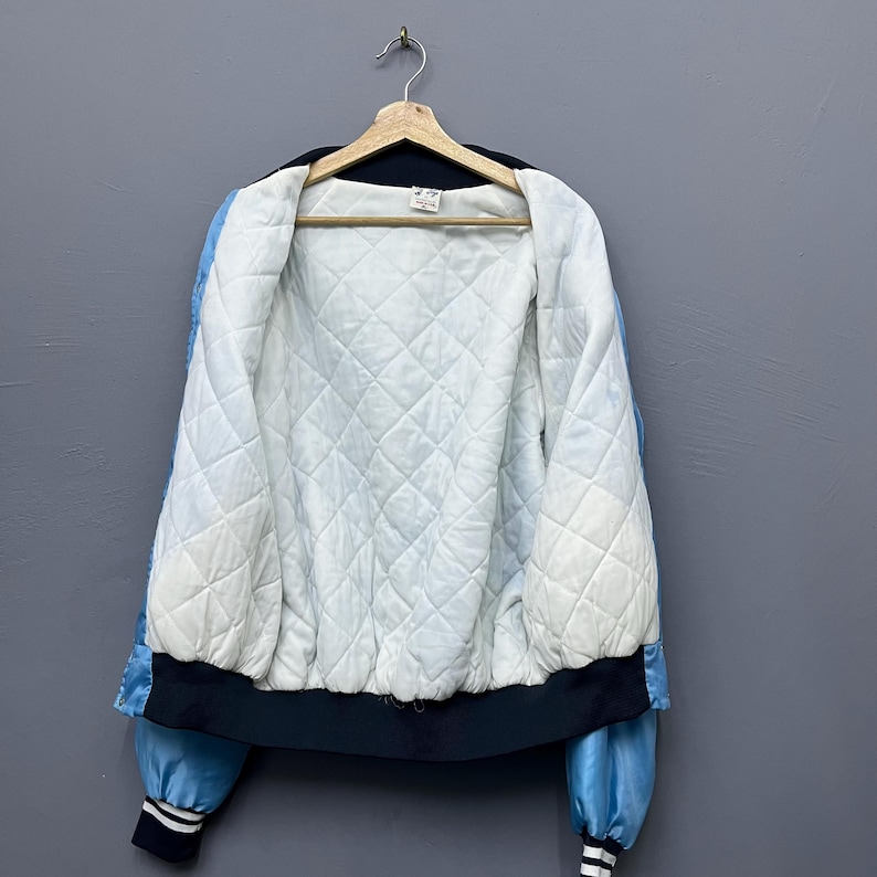 May include: A vintage satin bomber jacket in light blue and black, hanging on a wooden hanger. The jacket features a quilted white lining and black ribbed cuffs and hem. The sleeves have a light blue stripe.