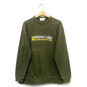 May include: Olive green crew neck sweatshirt with the text "DCSHOECOUSA A SKATEBOARD SHOE AND APPAREL COMPANY" in a yellow and black rectangular design. The sweatshirt is hanging on a wooden hanger.