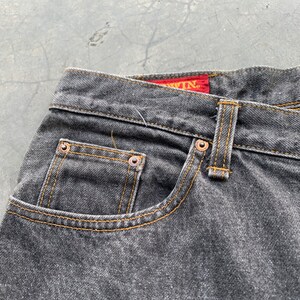 May include: Close-up of a pair of black denim jeans. The jeans feature a classic five-pocket design with copper rivets and a yellow stitched seam. The brand name is visible on a red tag.