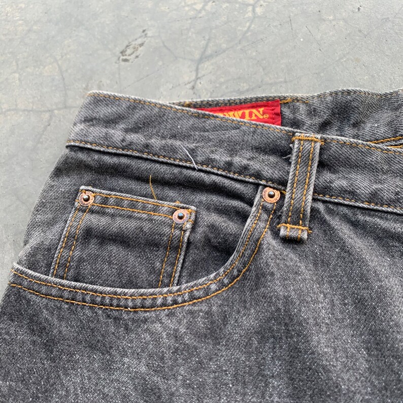 May include: Close-up of a pair of black denim jeans. The jeans feature a classic five-pocket design with copper rivets and a yellow stitched seam. The brand name is visible on a red tag.