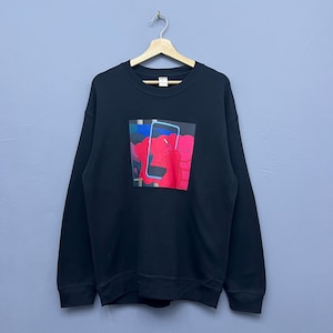 May include: A black long-sleeve crewneck sweatshirt featuring a central graphic print of a red cartoon character with X-shaped eyes holding a blue smartphone. The garment is displayed on a wooden hanger, showcasing a standard fit and ribbed cuffs.