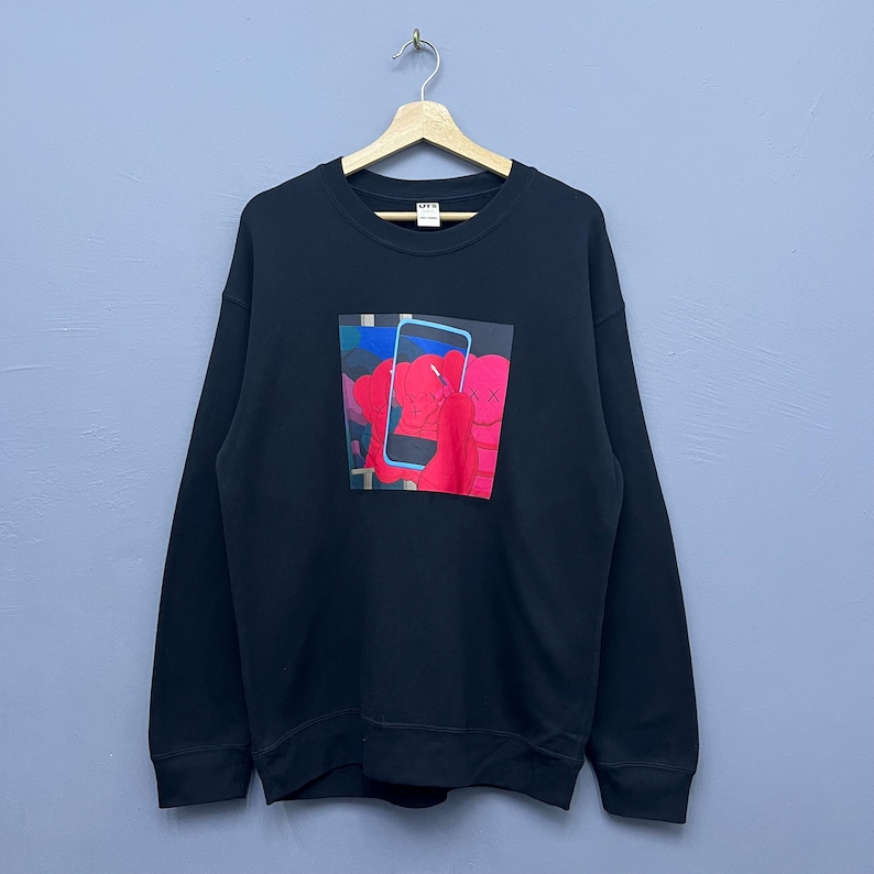 May include: A black long-sleeve crewneck sweatshirt featuring a central graphic print of a red cartoon character with X-shaped eyes holding a blue smartphone. The garment is displayed on a wooden hanger, showcasing a standard fit and ribbed cuffs.